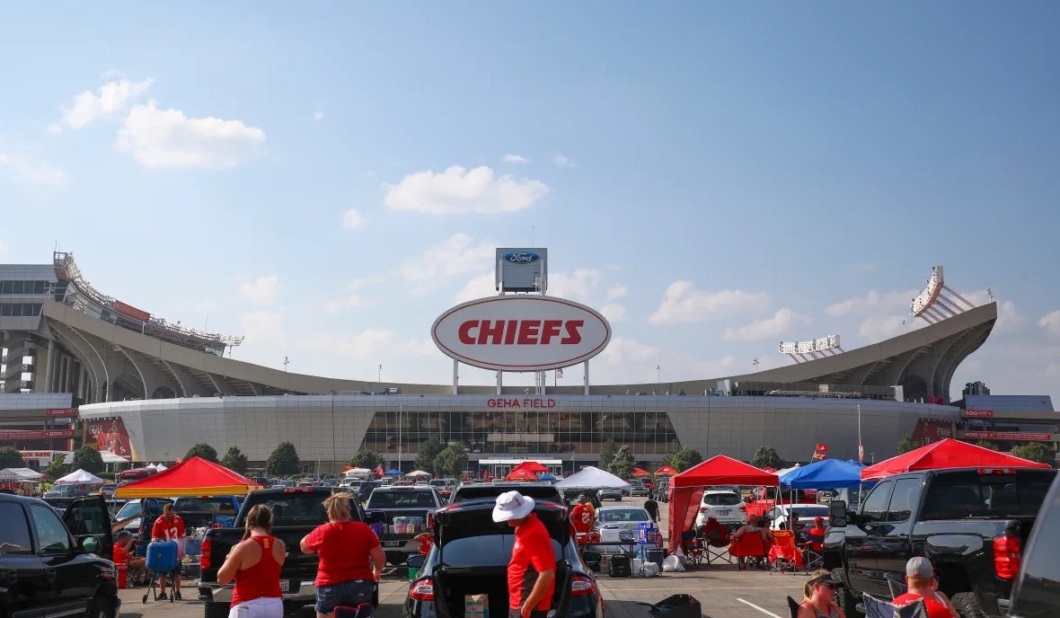 Chiefs fans tailgating in front of GEHA Field at Arrowhead Stadium on a clear, sunny day with a blue sky.